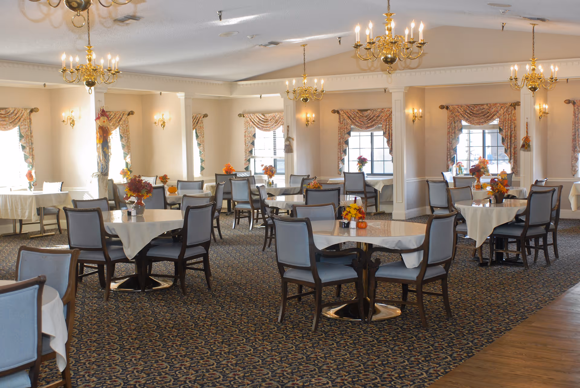 Dining room with multiple round tables set with white tablecloths, chairs, chandeliers, and floral centerpieces.