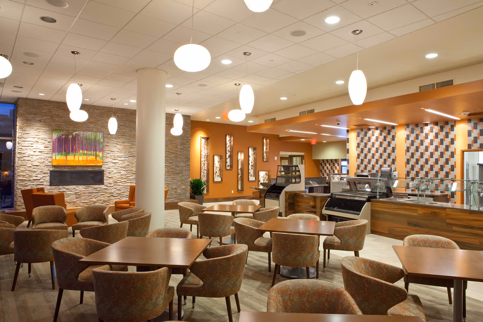 A spacious dining area with multiple tables and cushioned chairs arranged neatly. The room features warm lighting with hanging pendant lights, a stone accent wall with a colorful painting, and a serving counter with glass partitions. The decor includes patterned wallpaper and modern wall art, creating a welcoming atmosphere.