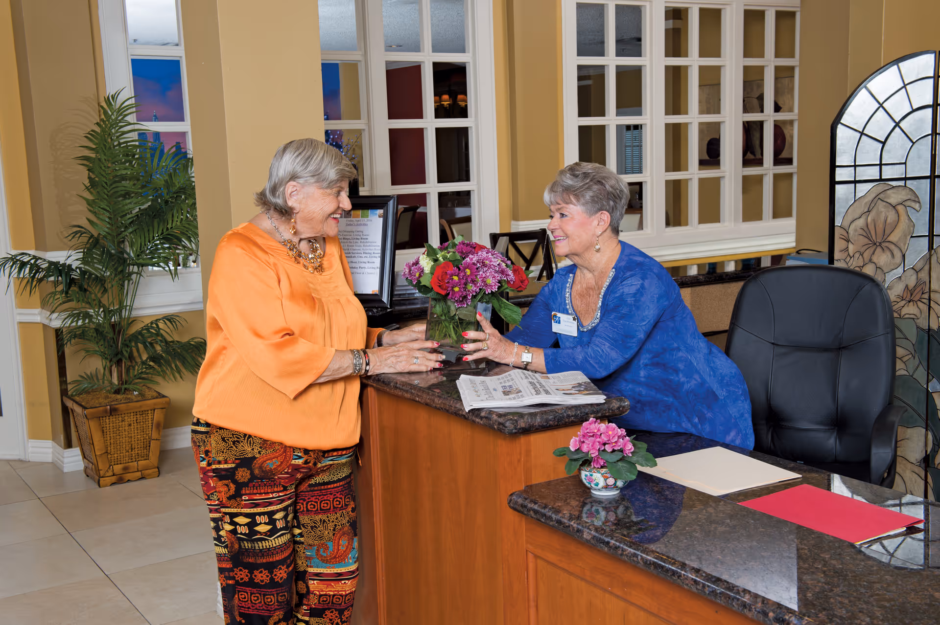 Two elderly women smiling and interacting at a reception desk in a warmly decorated interior space with plants and decorative windows.