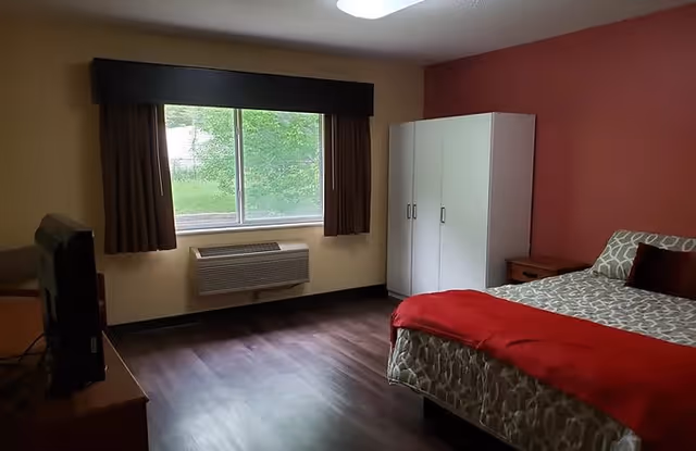 A simple bedroom with a bed covered in patterned bedding and a red blanket, a white wardrobe, a wooden nightstand, a window with brown curtains, and a television on a wooden stand. The walls are painted beige and red, and the floor is dark wood.