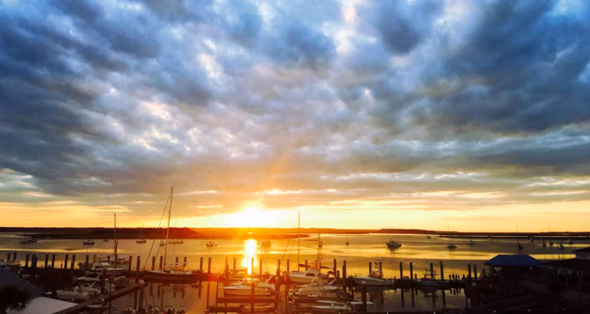 Sunset over a marina with boats and docks reflected on calm water beneath a dramatic cloudy sky.