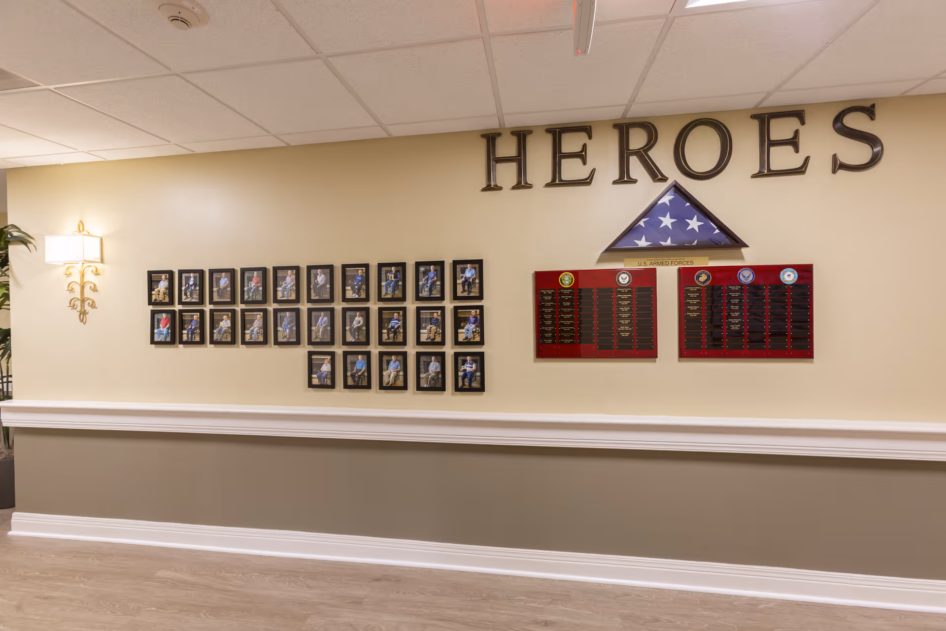 Wall display in a senior living facility hallway with the word 'HEROES' above a folded American flag and plaques honoring U.S. Armed Forces veterans, alongside framed photos of residents or veterans.