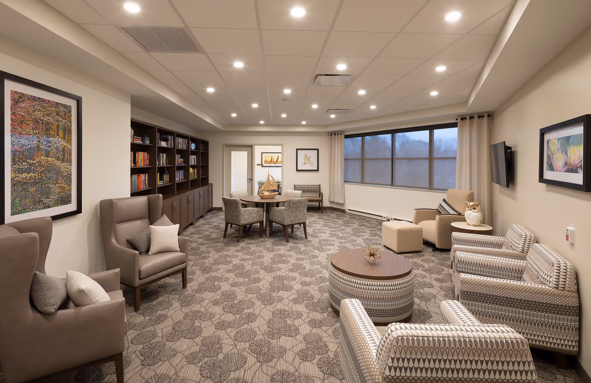 A spacious and well-lit living room area in a senior living facility with patterned carpet flooring, several upholstered armchairs and a round ottoman-style coffee table. There is a large window with curtains on one side, a wall-mounted TV, framed artwork on the walls, and a bookshelf filled with books along one wall. A round table with four chairs and a decorative sailboat centerpiece is positioned near the back of the room.