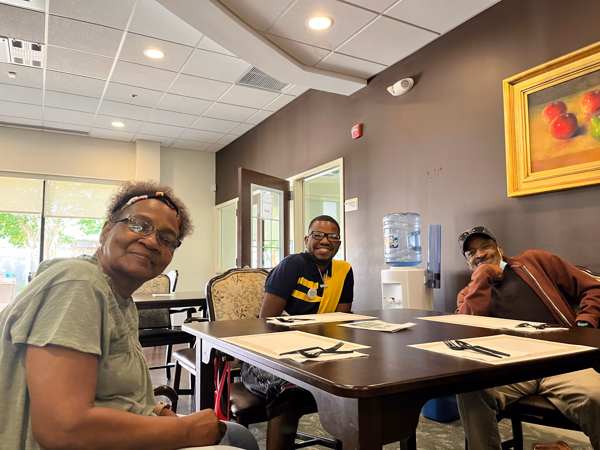 Three adults sitting around a dining table in a well-lit room. The table is set with placemats, napkins, and utensils. There is a water dispenser against a dark brown wall with a framed painting of fruit above it. Large windows with blinds allow natural light into the room.