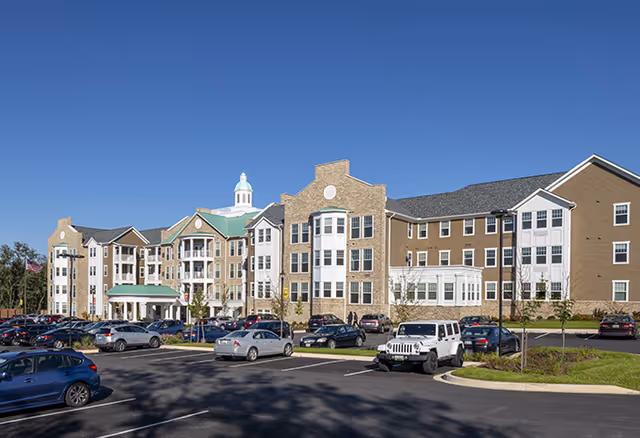 Exterior view of a large senior living facility building with multiple floors, a cupola on the roof, and a parking lot filled with cars in front under a clear blue sky.
