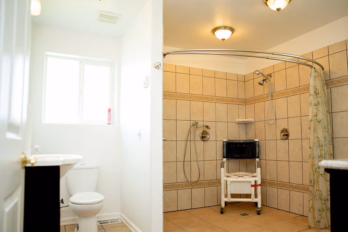Accessible tiled bathroom featuring a roll-in shower with a shower chair and handheld showerheads, plus a toilet and sink.