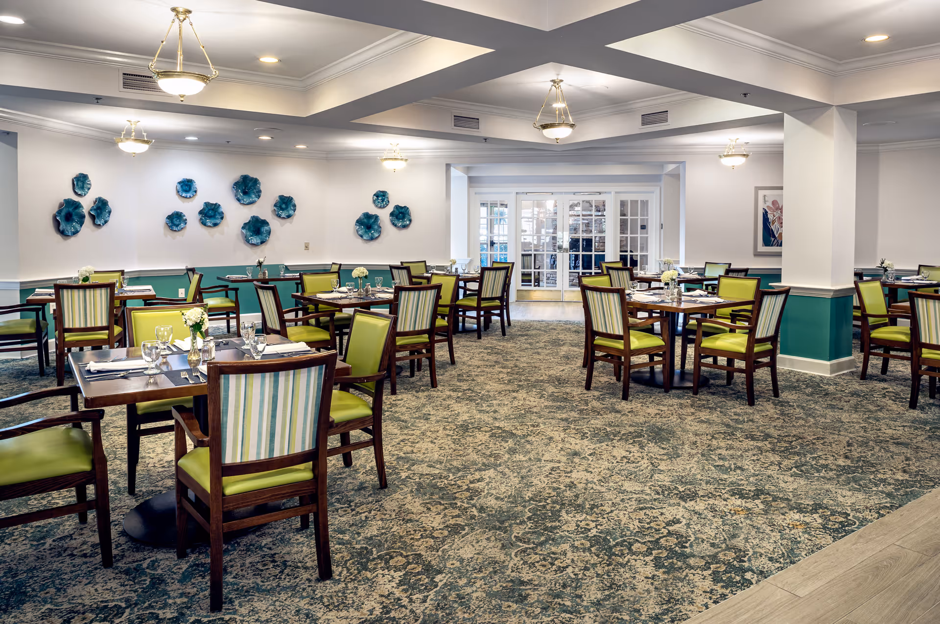 A spacious dining room with multiple wooden tables and chairs featuring green and striped upholstery. Each table is set with glassware, silverware, and small floral centerpieces. The room has patterned carpet flooring, white walls with teal wainscoting, decorative blue wall art, and ceiling light fixtures. Double glass doors are visible at the far end of the room.