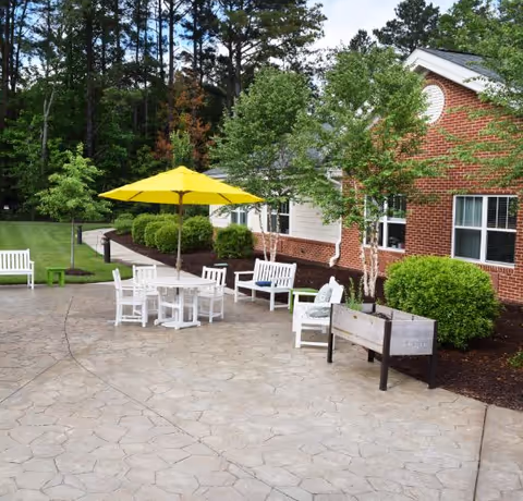 Outdoor patio area at Spring Arbor Cottage of Richmond featuring white chairs and tables with a yellow umbrella, surrounded by green bushes and trees next to a red brick building.