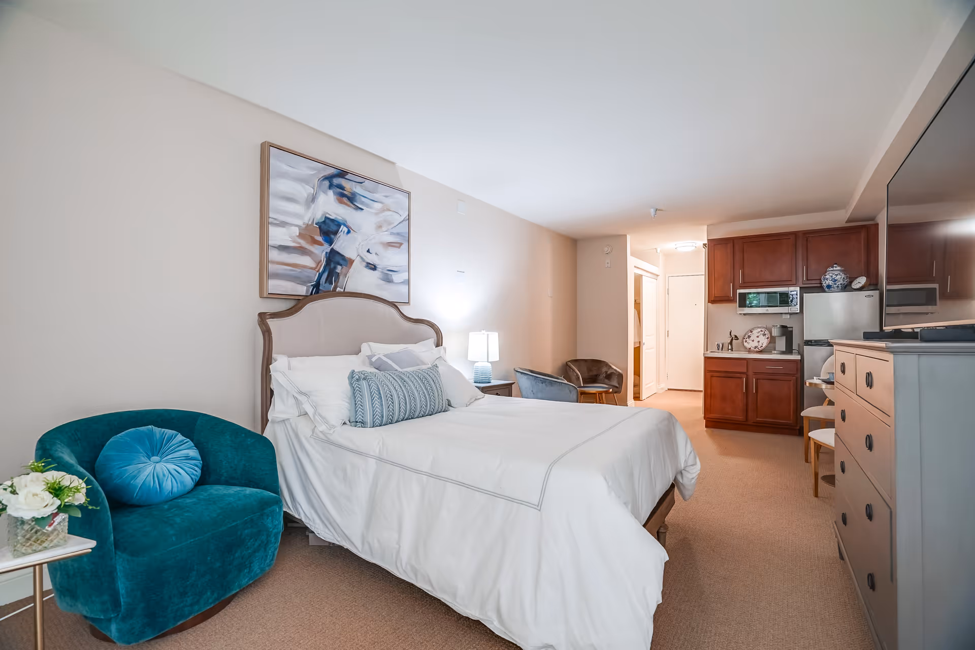 A furnished studio-style bedroom with a bed, teal armchair, dresser, and a small kitchenette in the background.