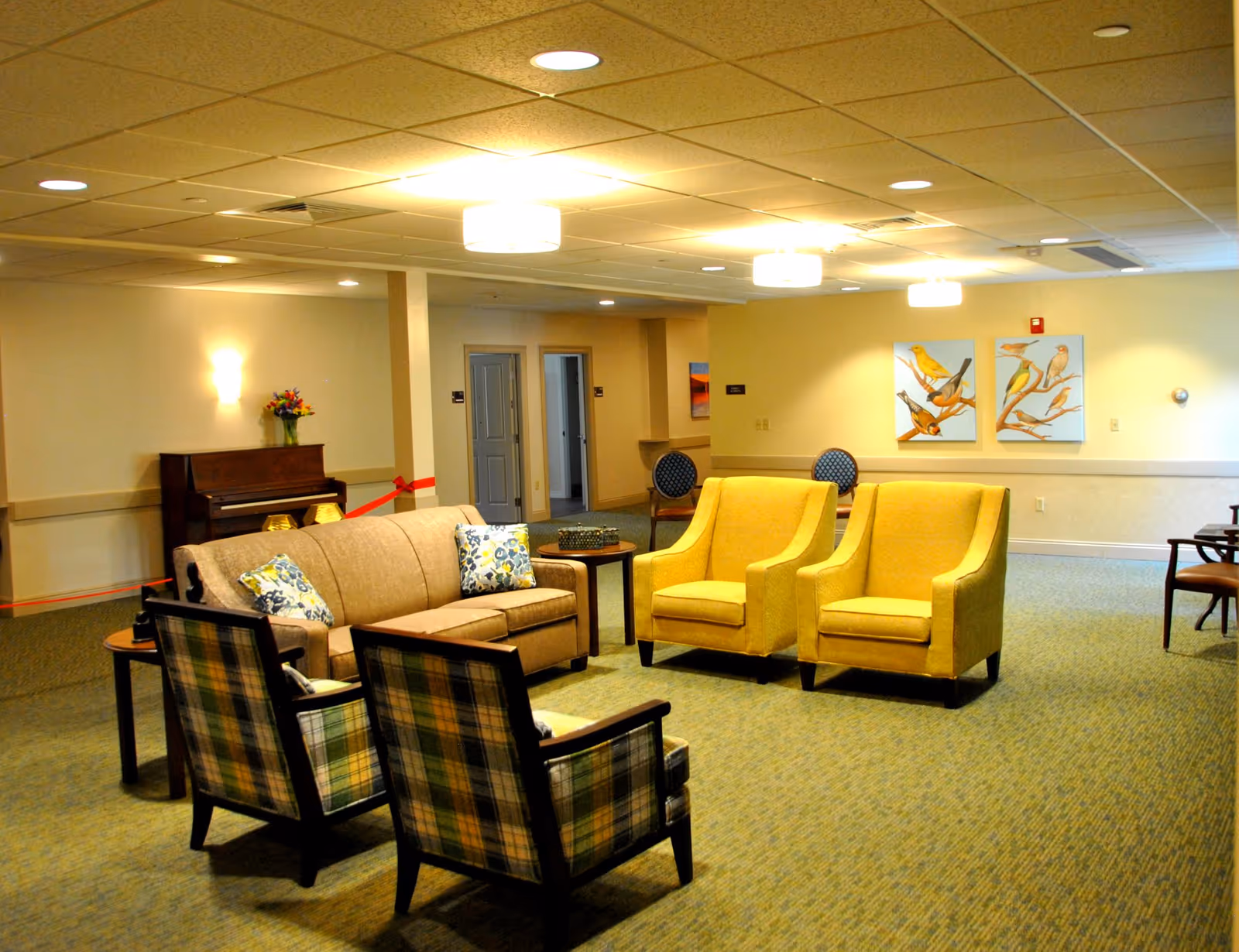 A well-lit communal lounge with sofas and armchairs arranged around tables, a piano in the background, and bird paintings on the wall.