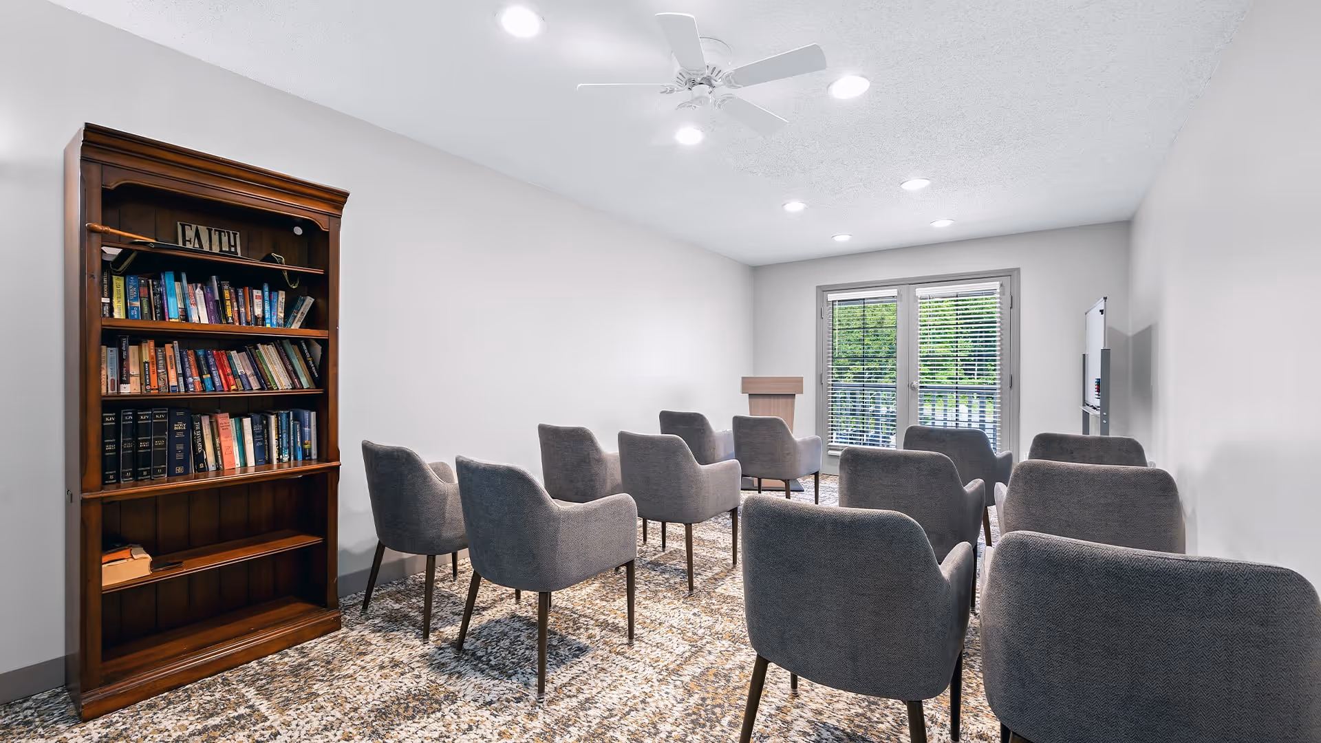 A small meeting or chapel room with gray upholstered chairs arranged in rows facing a wooden podium and a whiteboard. There is a wooden bookshelf filled with books on the left side of the room, and double glass doors with blinds at the far end letting in natural light.