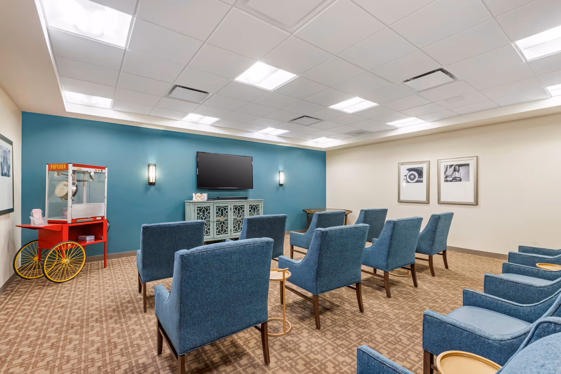 A small theater or media room with two rows of blue upholstered chairs facing a flat-screen TV mounted on a teal accent wall. There is a popcorn machine on a red cart with yellow wheels in the corner, and two framed black and white photos on the adjacent beige wall. The room has a patterned carpet and bright ceiling lights.