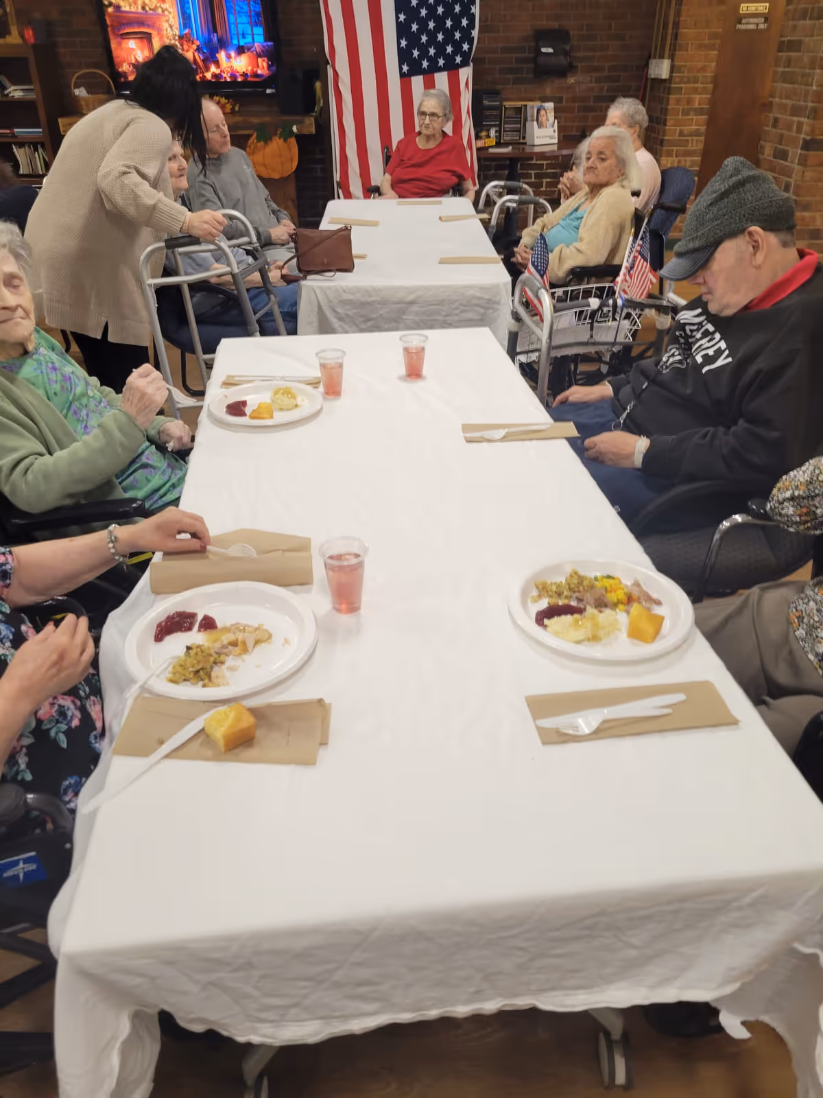 A group of elderly people seated around two tables covered with white tablecloths, having a meal with plates of food and cups of pink drink. A caregiver is assisting one of the elderly individuals. The room has a brick wall with an American flag hanging and a TV showing a fireplace scene in the background.