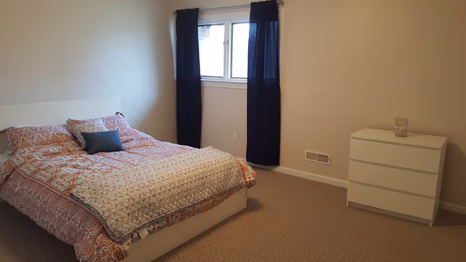 A simple bedroom with a bed covered in a patterned orange and beige comforter and pillows, a small white dresser with a glass candle holder on top, beige carpet, beige walls, and a window with dark blue curtains.