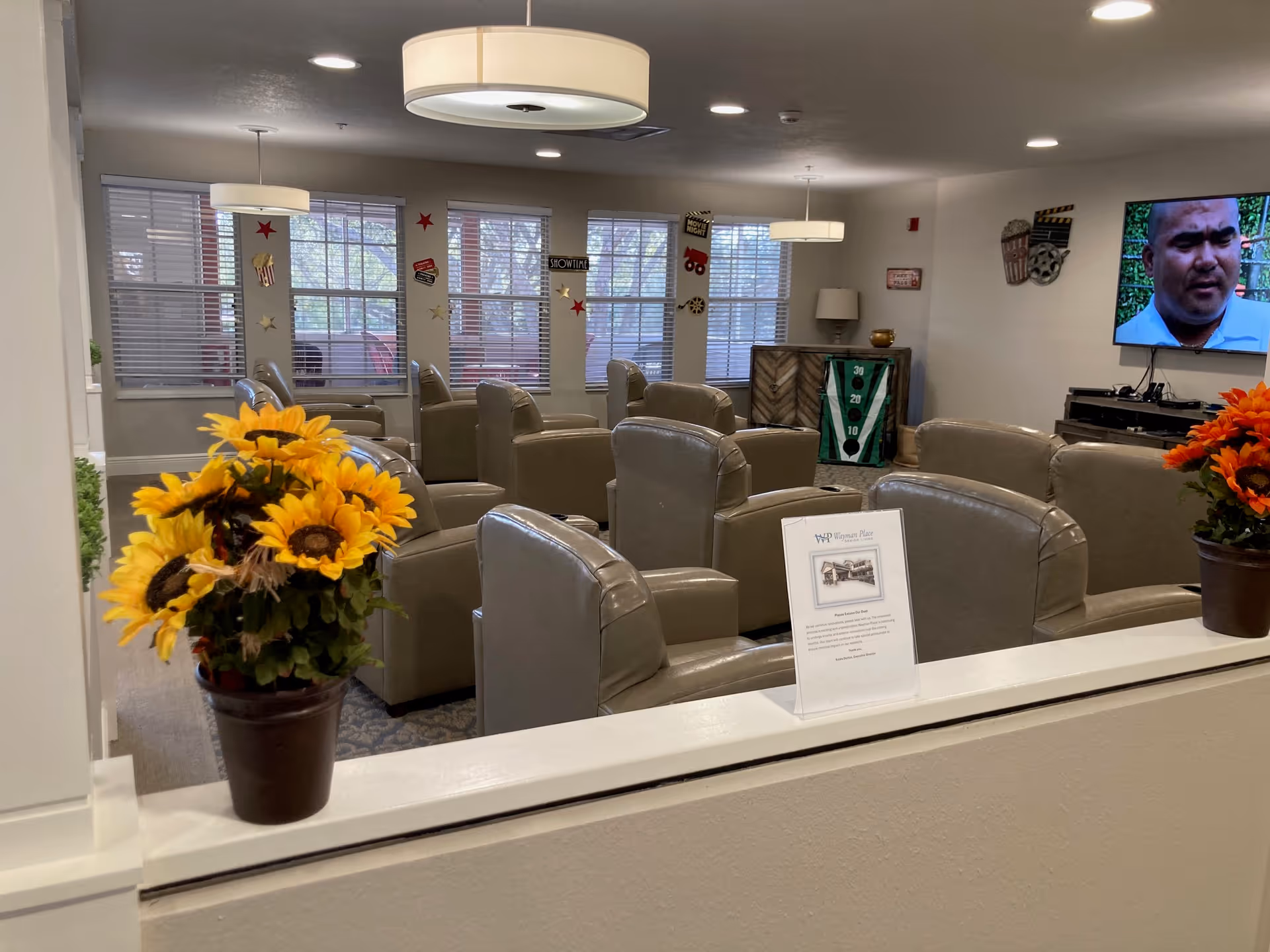 A senior living facility common area with multiple beige leather armchairs arranged in rows facing a wall-mounted television. The room has large windows with blinds, decorative movie-themed wall hangings, and two pots of sunflowers placed on a white half-wall in the foreground. Ceiling lights provide illumination.