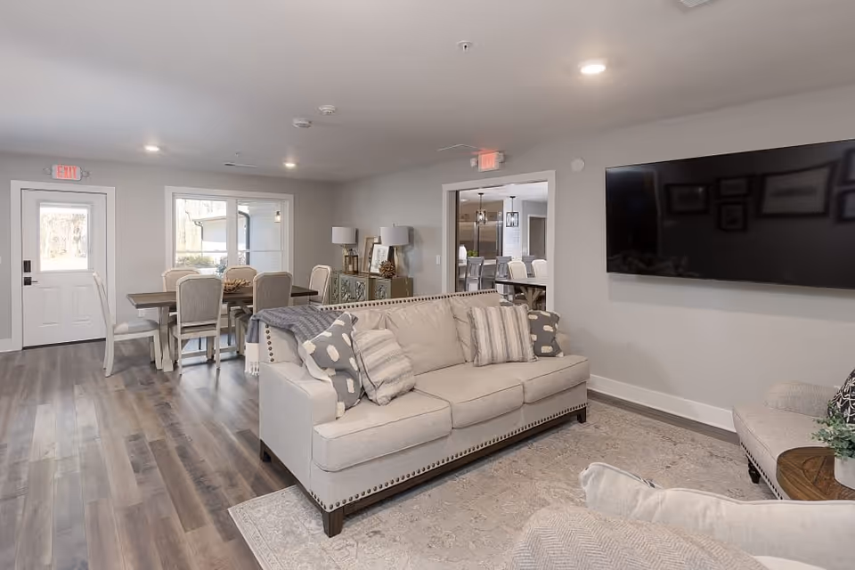 A bright and spacious senior living common area featuring a beige sofa with patterned cushions, a large wall-mounted flat-screen TV, and a dining table with six upholstered chairs near a door and window. The room has wood flooring, neutral-colored walls, and recessed ceiling lights. A doorway leads to another room with additional seating and a kitchen area visible in the background.