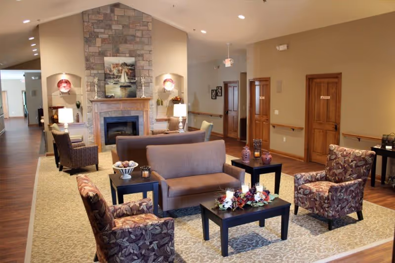A cozy senior living facility common area with a stone fireplace as the focal point. The room features a beige carpet with floral patterns, several upholstered chairs with floral and solid patterns, two small black coffee tables with decorative items, and warm lighting from table lamps. The walls are painted in a neutral tone with wooden doors and handrails along the hallway.