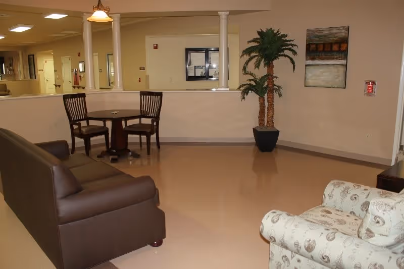 Interior view of a common area in a senior living facility with a brown leather sofa, a patterned armchair, a small round table with two wooden chairs, a potted artificial palm tree, and a painting on the wall.