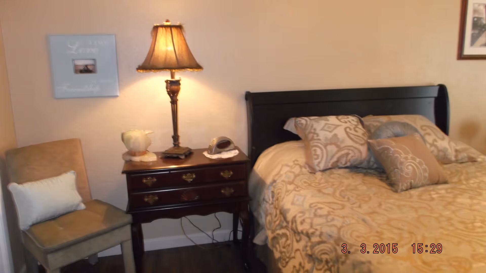 A cozy bedroom corner featuring a bed with patterned beige and brown bedding and multiple pillows. Next to the bed is a dark wooden nightstand with a decorative lamp, a small figurine, and a clock. A cushioned chair with a white pillow is placed beside the nightstand. The wall behind has a framed picture and a decorative sign that reads 'Live True Love Family'.