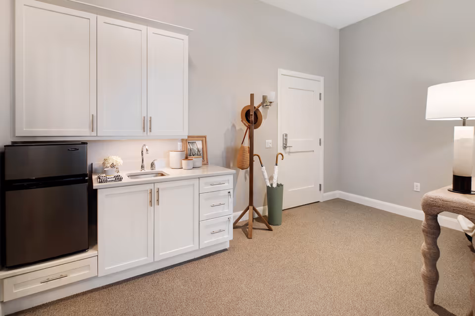 Carpeted studio room with white kitchenette cabinets and mini fridge, a coat rack with umbrellas and a floor lamp near a closed door.