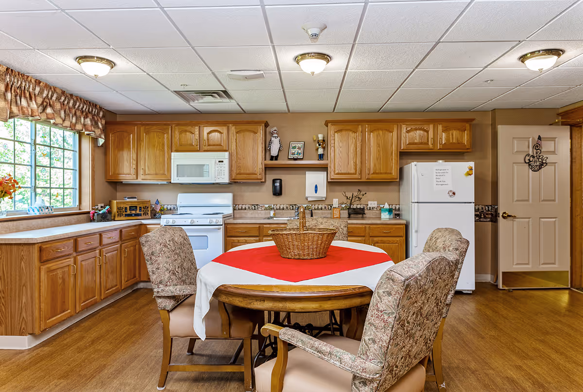 A cozy kitchen area in a senior living facility with wooden cabinets, a white microwave, stove, and refrigerator. There is a round dining table in the center covered with a white and red tablecloth, surrounded by four cushioned chairs with floral upholstery. A large window with patterned curtains lets in natural light, and the floor is wooden. The door on the right has a decorative hanging.