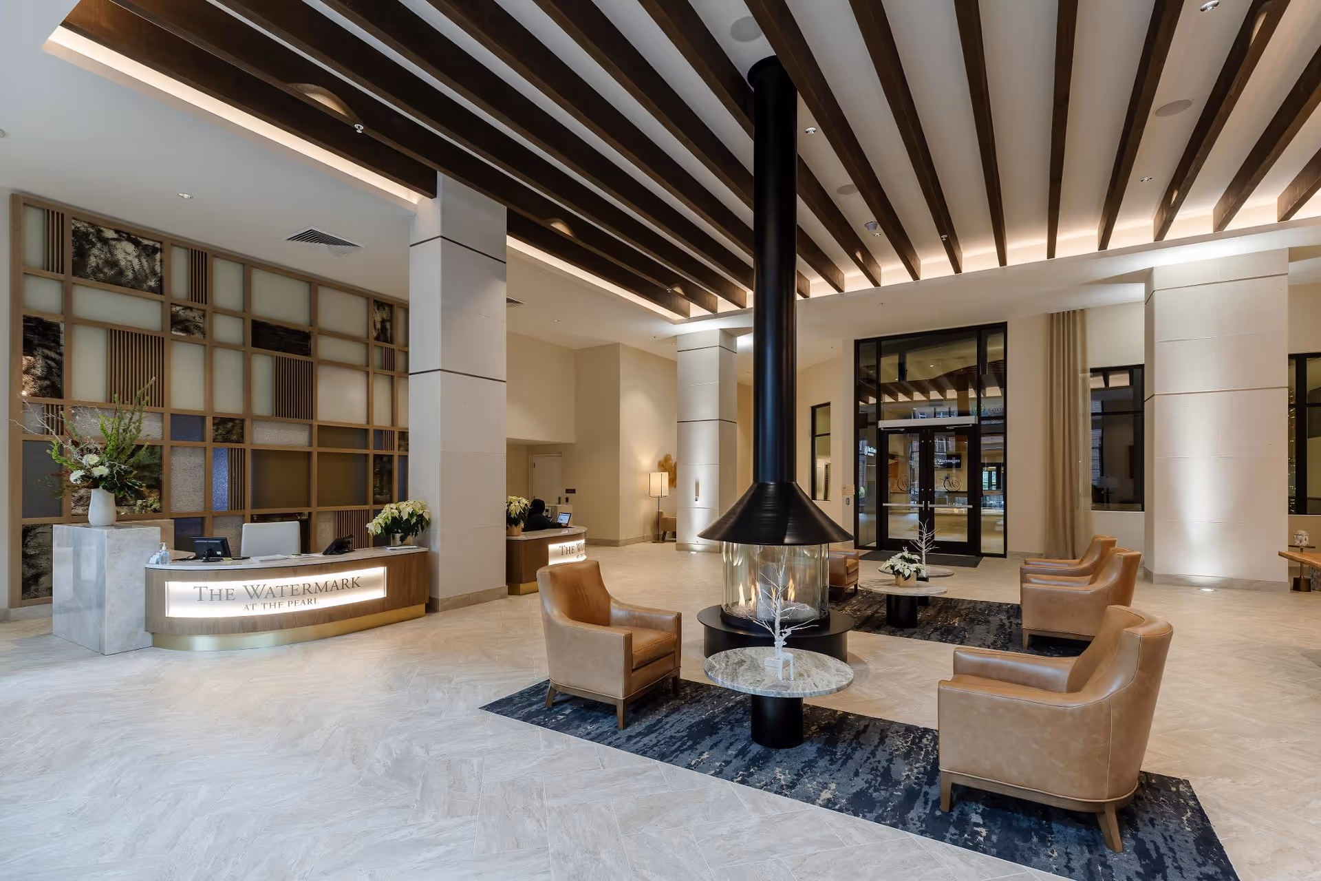Spacious and modern lobby area of The Watermark at the Pearl featuring a central suspended fireplace surrounded by four tan leather chairs and small round tables. The reception desk is visible on the left with a decorative wall behind it. The ceiling has wooden beams with recessed lighting, and large glass doors are at the far end of the room.