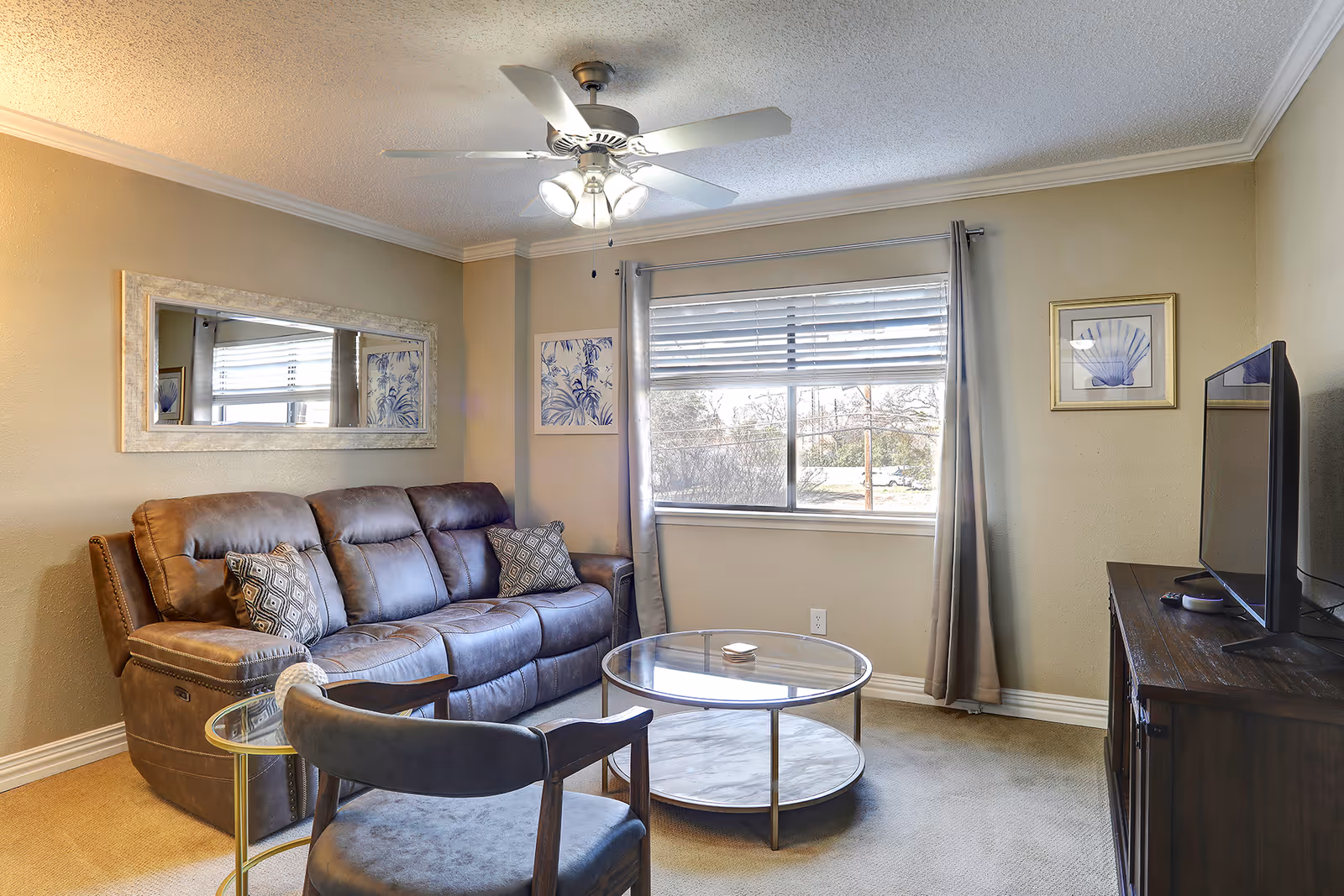 A cozy living room with a brown leather sofa, a gray cushioned chair, a round glass coffee table, and a wooden TV stand with a flat-screen television. The room has beige walls, a large window with blinds and gray curtains, a ceiling fan with lights, and framed artwork on the walls.