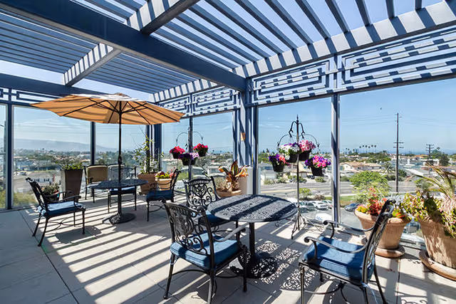 A bright outdoor patio area with a pergola casting striped shadows. The space features black metal tables and chairs with blue cushions, an orange umbrella, and several potted plants and hanging flower baskets. The patio overlooks a suburban area with houses and trees under a clear blue sky.