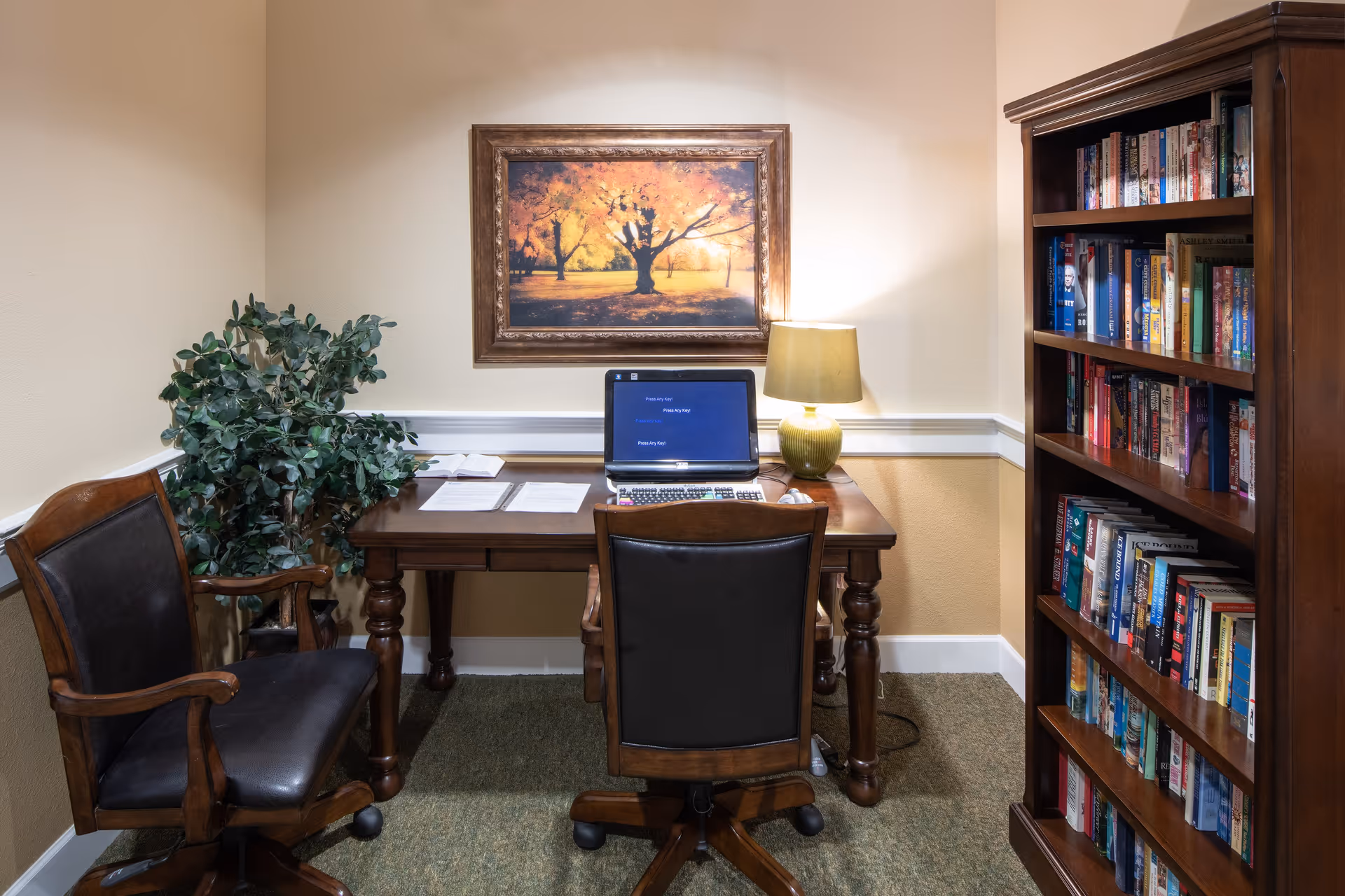A cozy office or study area with a wooden desk and two chairs, one facing the desk and the other to the side. On the desk is a laptop, some papers, a calculator, and a green table lamp. A framed picture of a tree with autumn leaves hangs on the wall above the desk. To the right is a wooden bookshelf filled with books, and to the left is a green leafy plant. The room has beige walls and carpeted flooring.