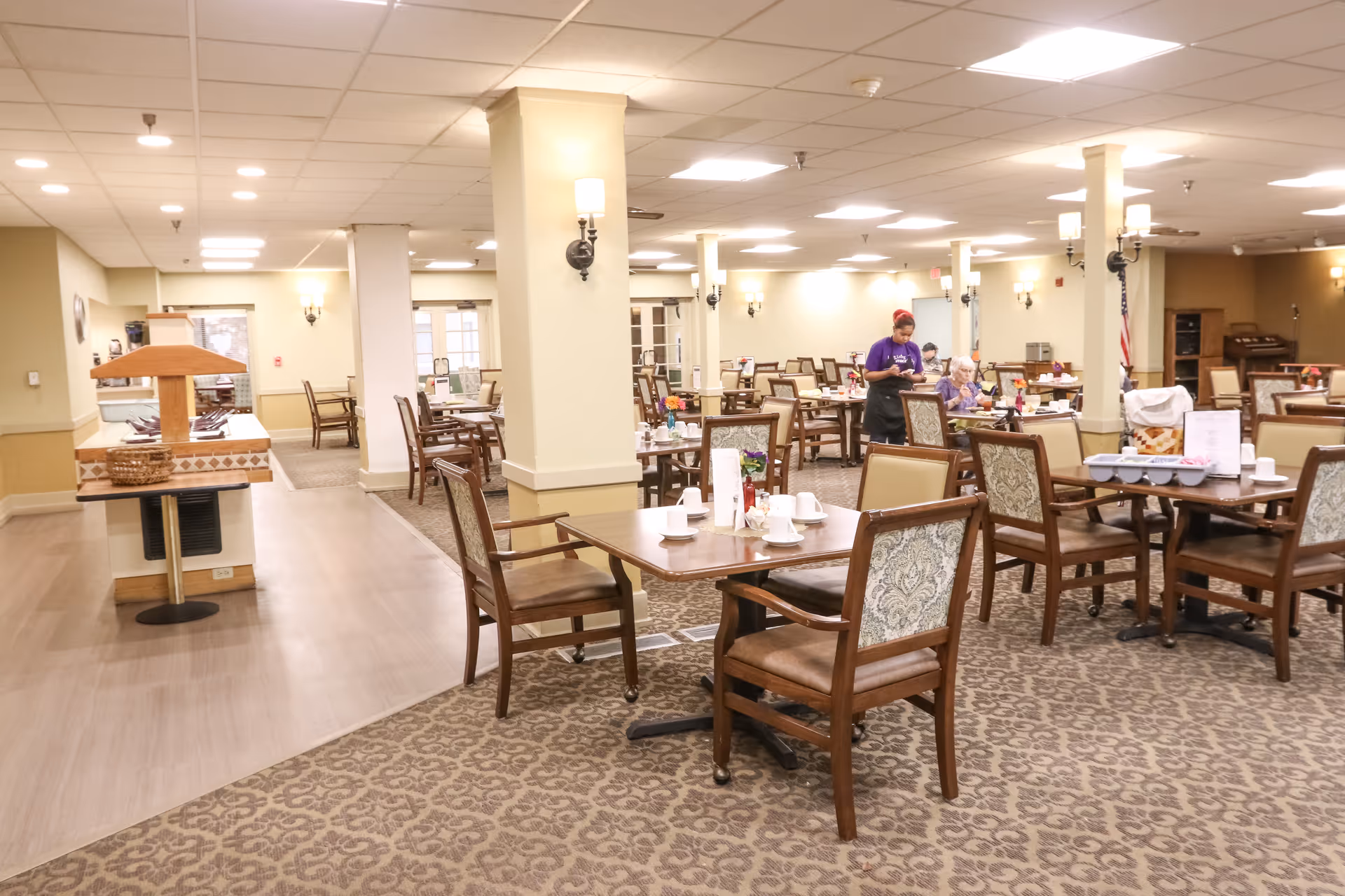 A spacious dining room with multiple wooden tables and chairs set for meals and staff assisting residents in the background.