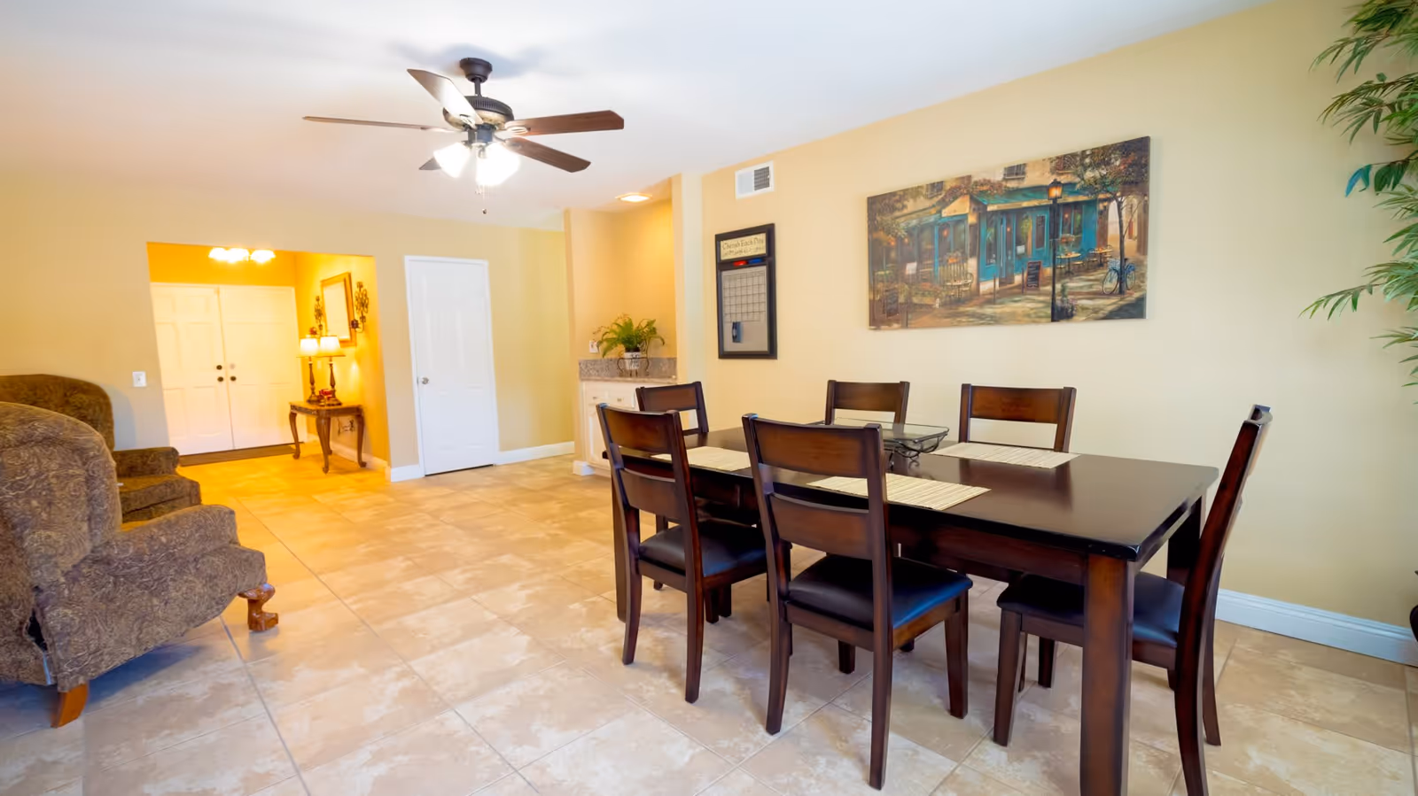 Bright communal dining area with a dark wood table and six chairs, tiled floor, ceiling fan and adjacent upholstered seating.