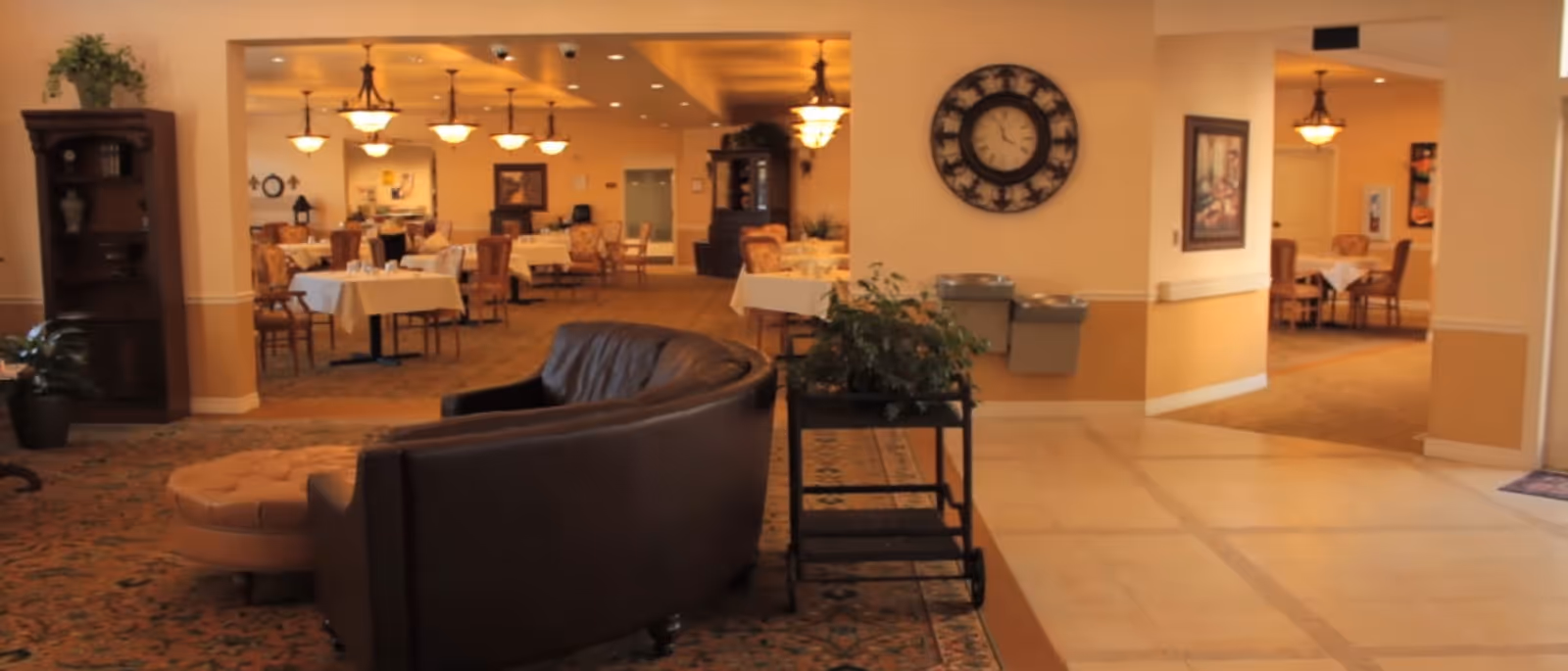 Interior view of a senior living facility showing a common area with a curved brown leather sofa and a round ottoman in the foreground, a plant on a small table, and a dining area with tables and chairs set with white tablecloths in the background. The walls are painted beige with decorative elements including a large wall clock and framed artwork.