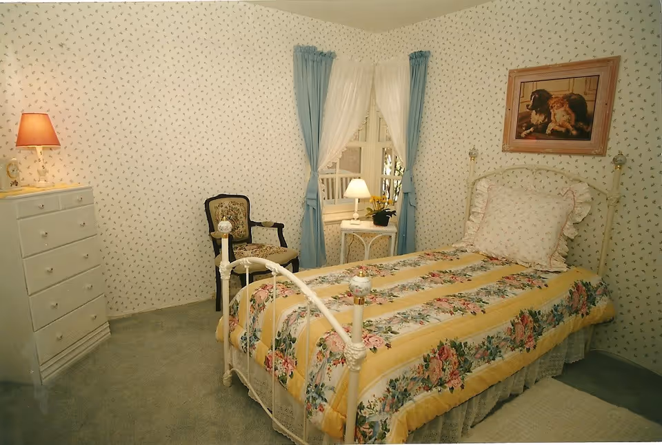 A cozy bedroom with a single bed featuring a floral and yellow striped bedspread and a ruffled pillow. The room has wallpaper with a small floral pattern, a white dresser with a lamp on top, a vintage upholstered chair, and a small white side table with a lamp and a potted plant near a window with blue and white curtains. A framed picture of dogs hangs on the wall above the bed.