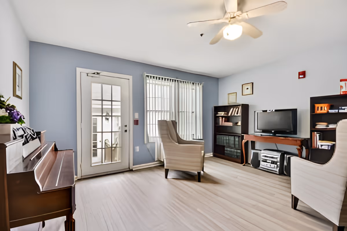 A bright living room with light wood flooring, light blue walls, and a white ceiling fan. The room features a wooden piano on the left, two beige armchairs, a TV on a wooden console table, and two dark wood bookshelves filled with books and decorative items. A glass door and a large window with vertical blinds allow natural light to enter the room.