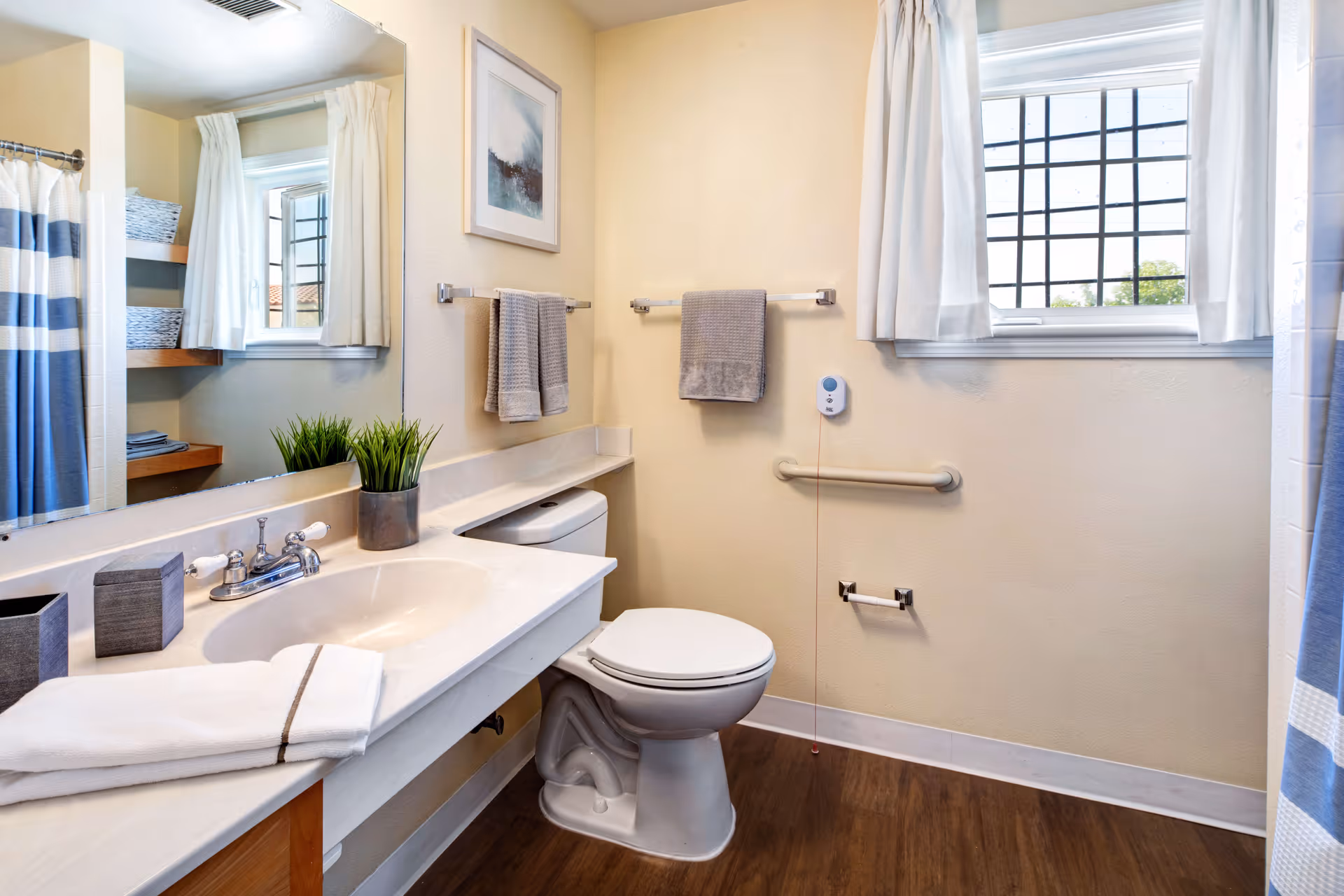 A clean and well-lit bathroom featuring a white sink with a silver faucet, a toilet, and a window with white curtains. There are two gray towels hanging on towel racks, a small potted plant on the counter, and a shower curtain with blue and white stripes. The floor is wood-style, and the walls are painted a light beige color.