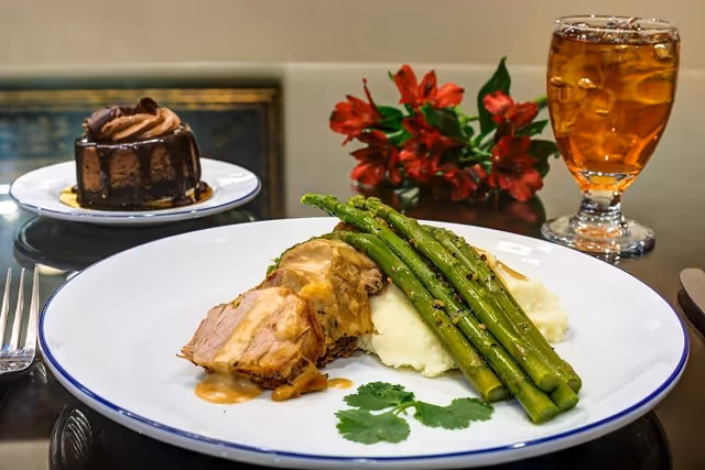 A plated meal consisting of sliced roasted meat with gravy, mashed potatoes, and grilled asparagus, garnished with a sprig of cilantro. In the background, there is a glass of iced tea, a small chocolate dessert on a white plate, and a red flower arrangement.
