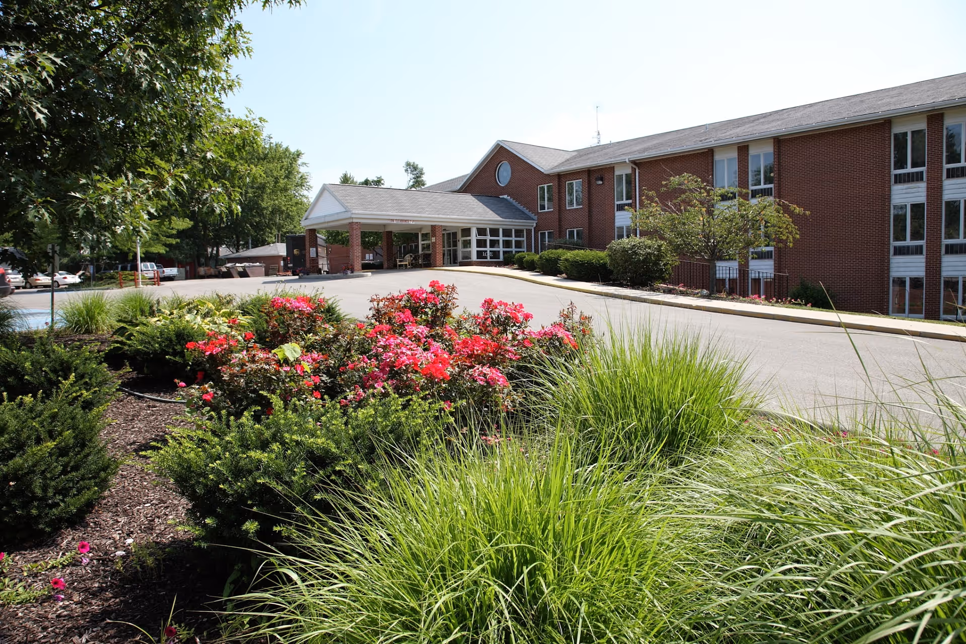 Entrance of a red-brick senior living facility with a covered drop-off, driveway and landscaped flower beds in the foreground.