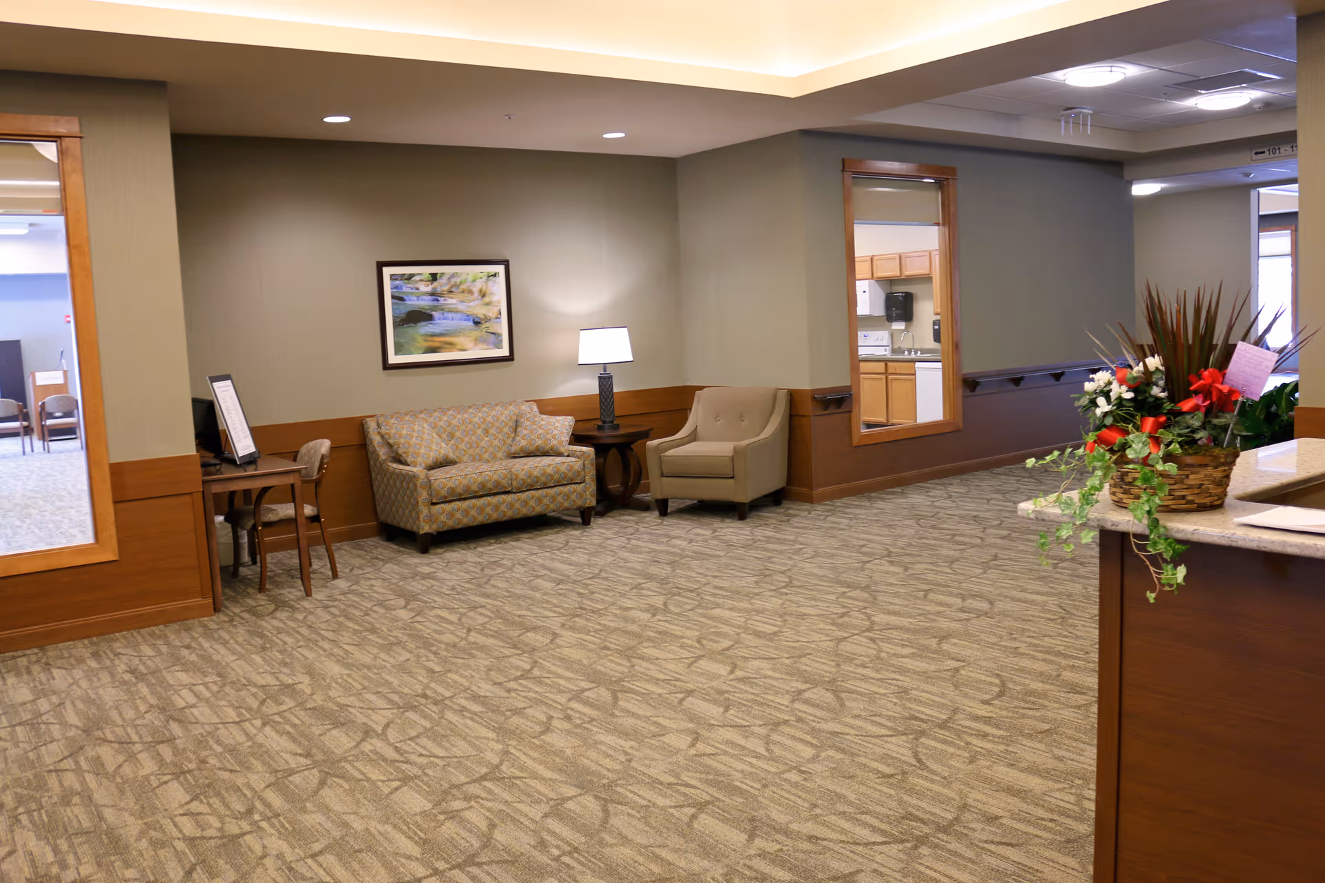 A spacious seating area in a senior living facility lobby with sofas, chairs, a lamp, framed art, and a floral arrangement on a reception counter.