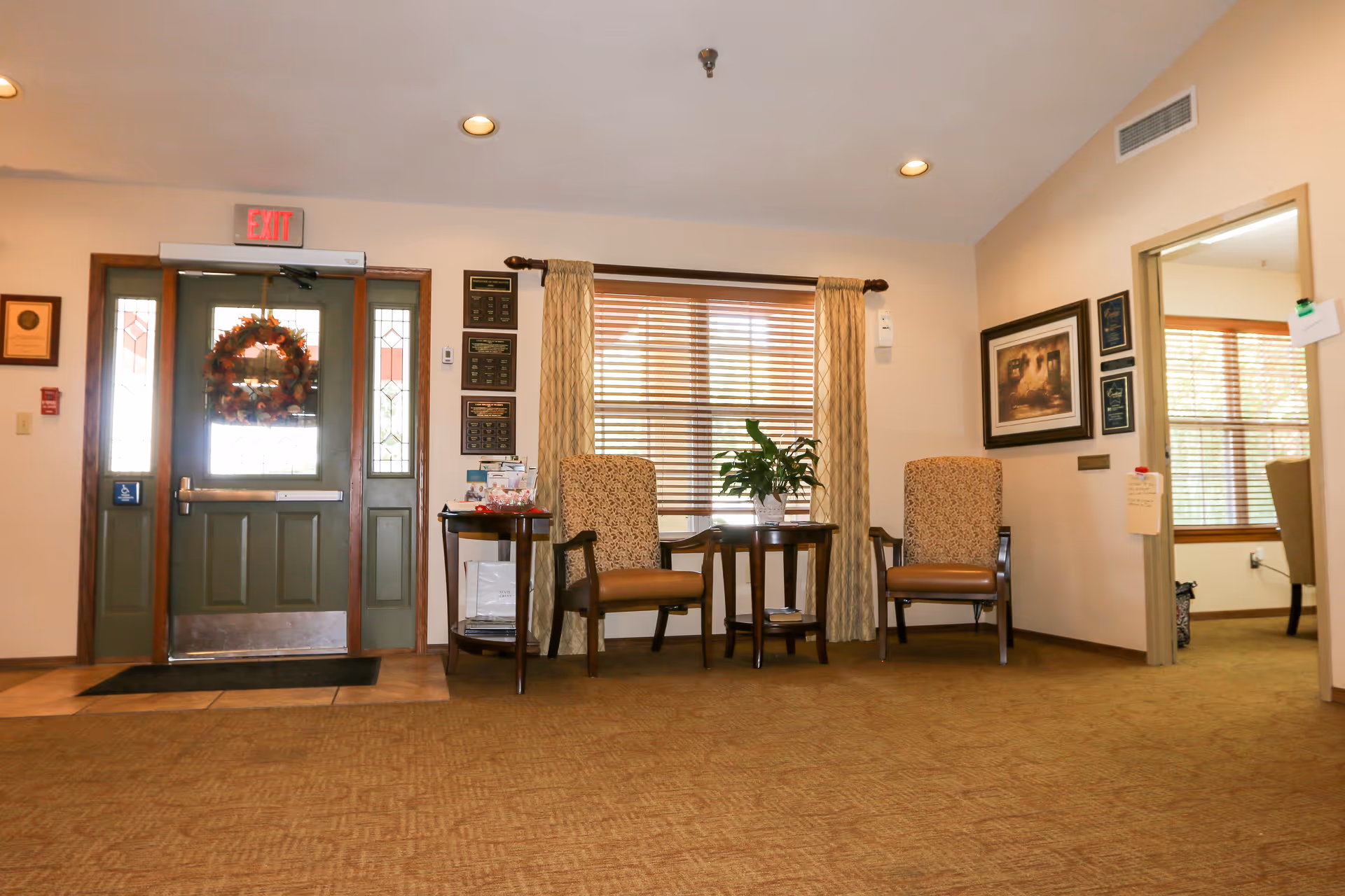 Interior view of a senior living facility entrance area with a green door decorated with a fall-themed wreath, two upholstered chairs with wooden arms, a small table with a plant, window with blinds and curtains, and framed pictures on the walls.