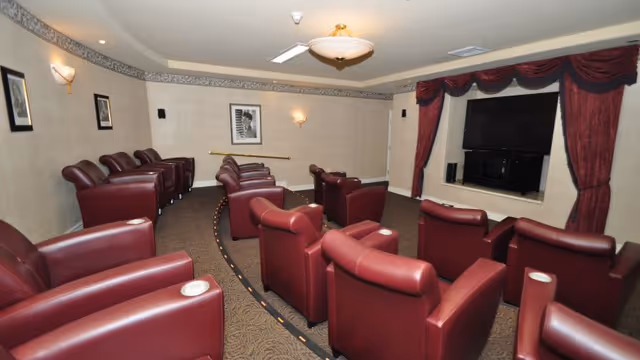 Small theater-style room with rows of red leather recliners facing a wall-mounted TV framed by red curtains.