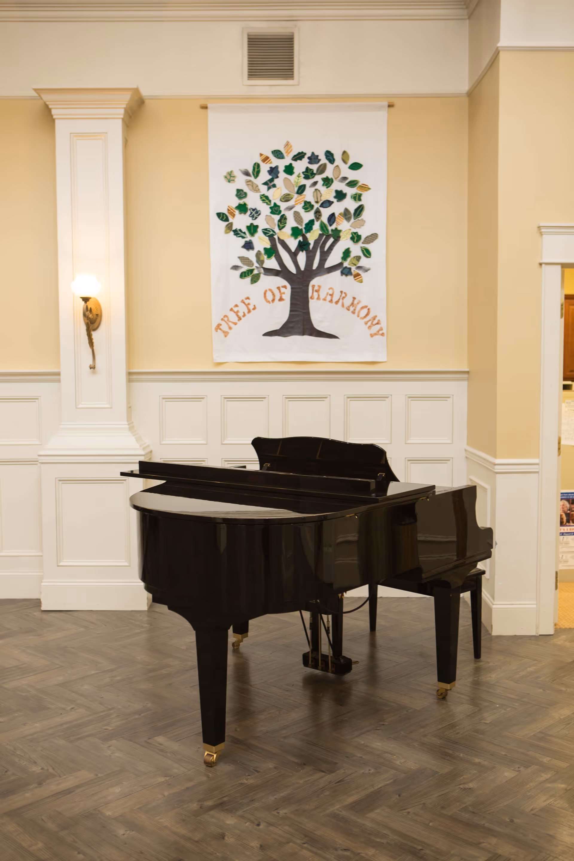 A black grand piano placed on a wooden floor in a room with cream-colored walls and white wainscoting. Above the piano, there is a decorative wall hanging featuring a tree with green and brown leaves and the words 'TREE OF HARMONY'. A wall sconce with a lit bulb is mounted on the left side of the wall.