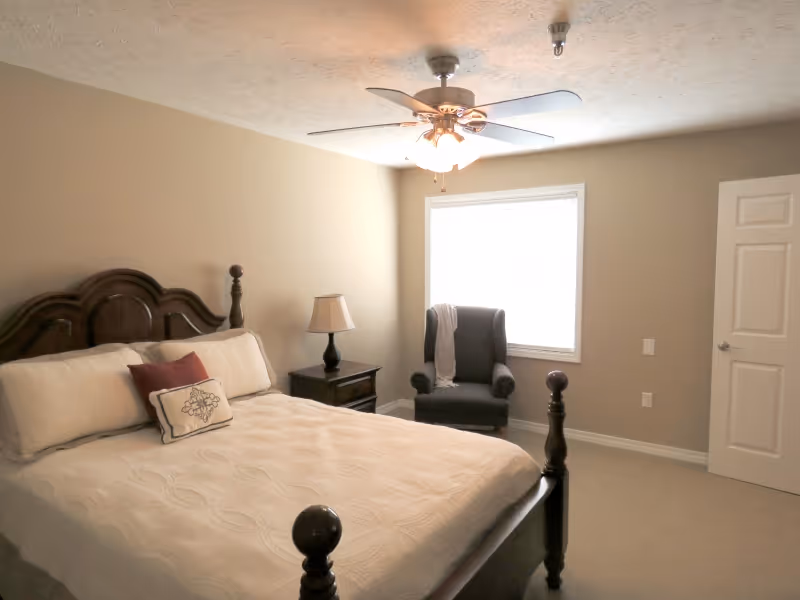 A bedroom with a large wooden bed featuring white bedding and decorative pillows. Next to the bed is a dark wooden nightstand with a lamp. A dark upholstered armchair with a light-colored throw is positioned near a window with white blinds. The room has beige walls, a white ceiling with a ceiling fan and light, and a white door slightly ajar.