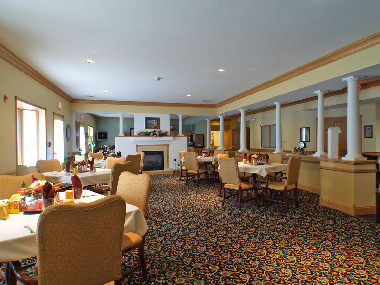 A spacious dining room in an assisted living facility with several round tables covered with white tablecloths, each set with chairs, napkins, and condiments. The room features a patterned carpet, a fireplace in the center, and decorative columns along one side. Large windows allow natural light to brighten the space.