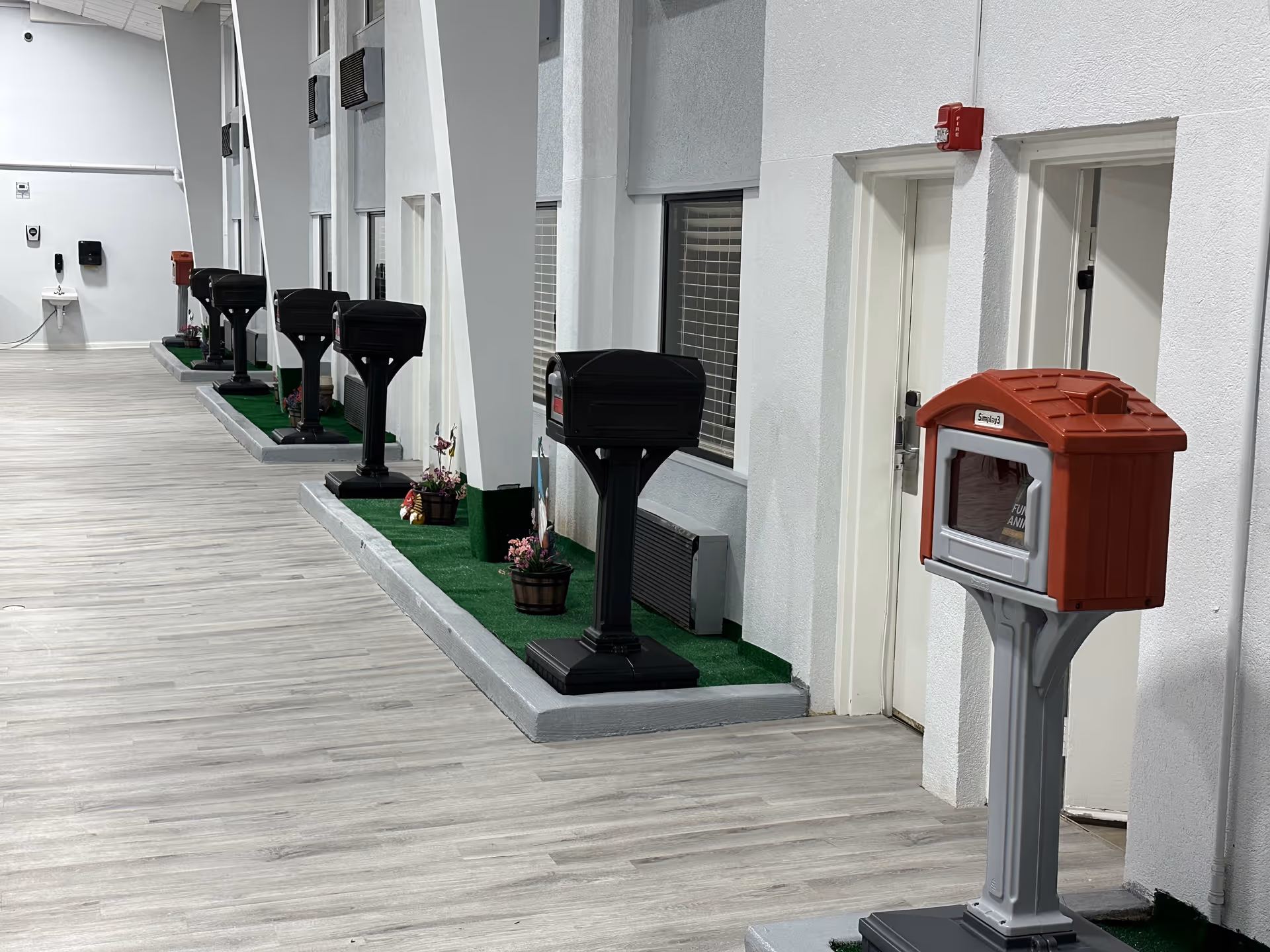 Indoor hallway with a row of black mailboxes mounted on stands along the wall, with small flower pots placed between them. At the front right, there is a red and gray mailbox on a stand. The floor is light wood, and the walls are white with windows and doors.