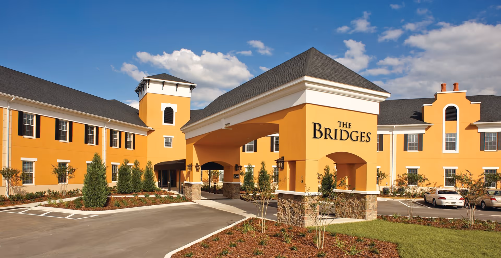 Exterior view of Sweet Water Grand The Bridges Assisted Living facility with a large yellow building featuring multiple windows, a covered entrance with stone pillars, a parking lot with cars, and a clear blue sky with some clouds.