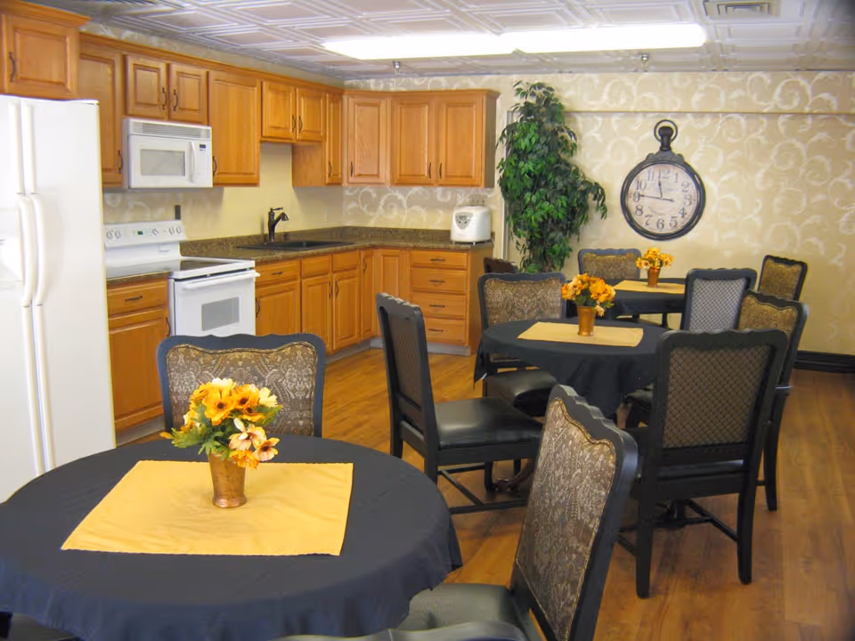 A dining area with round tables covered in black tablecloths and yellow placemats, each with a small vase of yellow flowers. The room has wooden flooring, a kitchen area with wooden cabinets, a white refrigerator, stove, microwave, and a countertop with a rice cooker. A large wall clock and a tall green plant are visible on the patterned wallpapered wall.