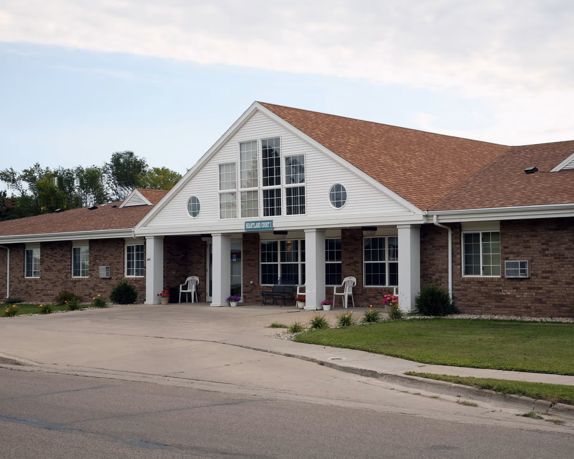 Exterior front view of a single-story brick building with a white gabled entrance and multiple windows. The entrance has white columns, a bench, plastic chairs, and flower pots. A sign above the entrance reads 'HEARTLAND COURT 1'. The building is surrounded by a paved driveway and a small grassy area with plants.