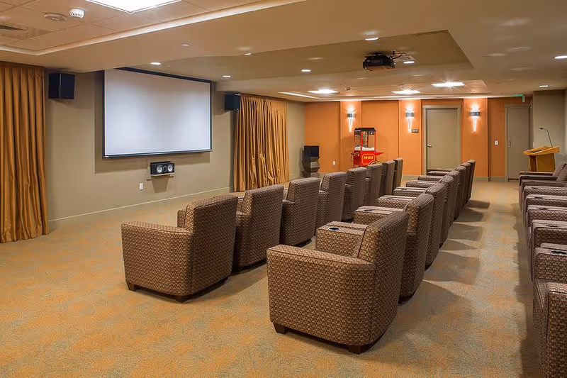 A small theater room with multiple rows of cushioned armchairs facing a large blank projection screen. The room has beige walls, orange accent walls, and brown curtains. There is a popcorn machine and a wooden podium at the back of the room.