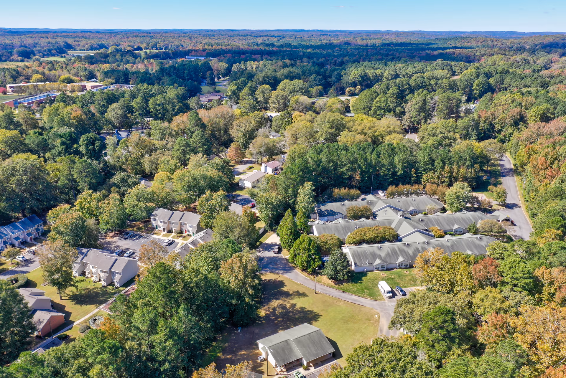 Aerial view of Cambridge Hills Assisted Living facility surrounded by dense trees and greenery, with multiple buildings and parking areas visible under a clear blue sky.