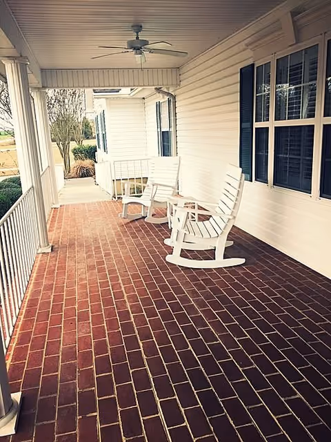 A covered porch with red brick flooring featuring two white wooden rocking chairs and a small round table between them. The porch has white railings and columns, a ceiling fan, and windows with dark shutters on the right side. Trees and bushes are visible in the background.